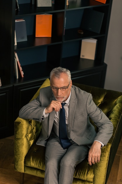 Thoughtful serious grey-haired man sitting in an olive green armchair while propping his chin