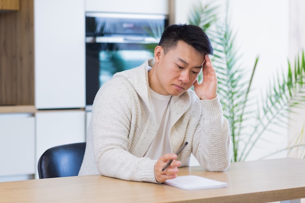 thoughtful serious young asian man looking away student writer sit at home office desk