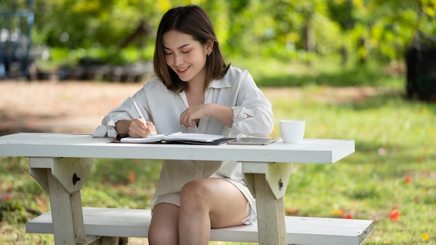 Задумчивая улыбающаяся женщина в парке, использующая блокнот и пишущая Портрет молодой очаровательной деловой женщины, проверяющей онлайн Деловая работа на своем смартфоне на открытом воздухе в парке на мягком зеленом фоне