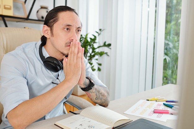 Thoughtful stressed graphic designer putting hands in pray gesture thinking about problem solution when looking at computer screen