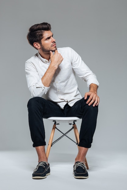 Thoughtful stylish attractive casual young man sitting on chair over gray wall