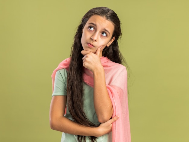 Задумчивая девочка-подросток в шали смотрит вверх, держа руку на подбородке, изолированную на оливково-зеленой стене с копией пространства