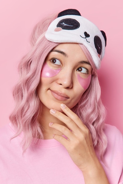 Thoughtful teenage girl with wavy pink dyed hair keeps hand on chin looks away applies hydrogel patches under eyes for skin treatment wears blindfold on forehead isolated over pink background