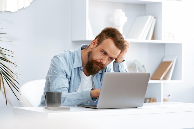 Thoughtful tired young bearded man sitting at the table working on laptop