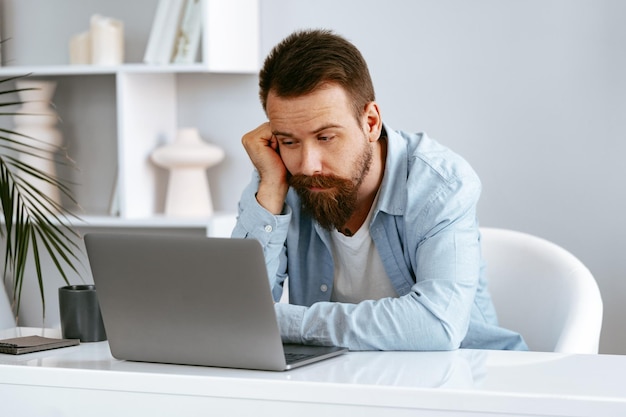 Thoughtful tired young bearded man sitting at the table working on laptop