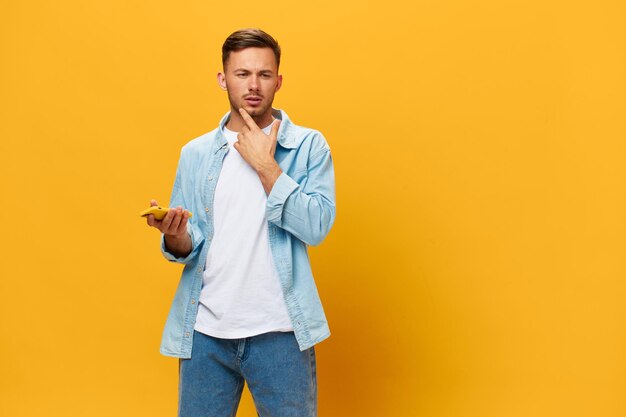 Thoughtful unhappy pensive handsome man in blue basic tshirt\
make difficult decision hold phone posing isolated on yellow studio\
background copy space banner mockup online social media\
concept