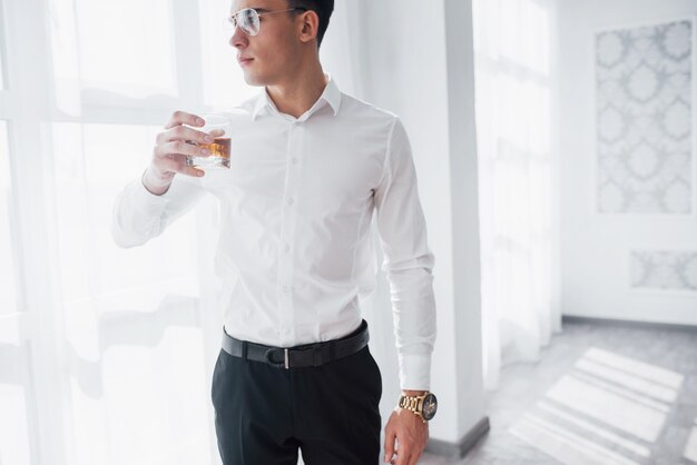 Thoughtful view. Gorgeous man in eyeglasses stand indoor and drink alcohol.