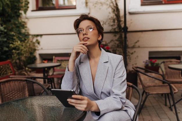 La donna premurosa in abito grigio tiene il tablet e si siede nel ristorante di strada l'affascinante signora dai capelli corti con una giacca elegante riposa nel caffè esterno