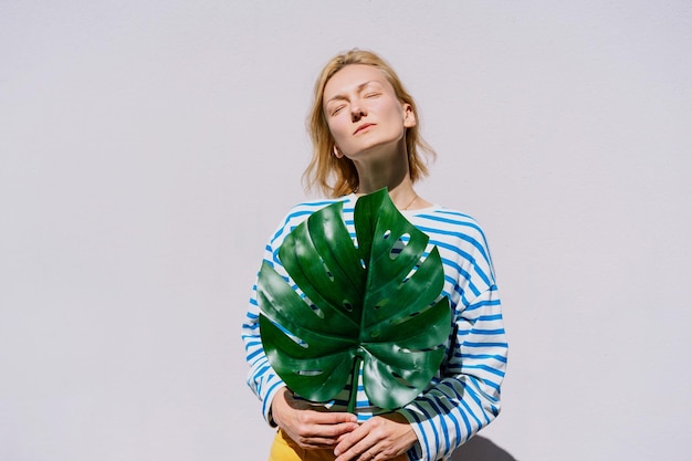 Thoughtful woman holding monstera leaf in front of wall Concept of Ecology Individuality Idea