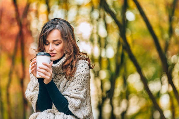 ニットのセーターを着た思いやりのある女性は、秋の公園でホットコーヒーを保持しています