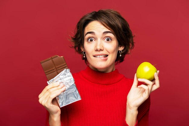 Foto donna pensierosa che tiene pensierosa una barretta di cioccolato e una mela che sceglie