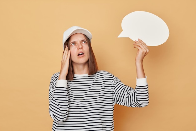 Thoughtful woman wearing baseball cap and striped shirt holding empty bubble speech cloud isolated over beige background being deep in thoughts thinking