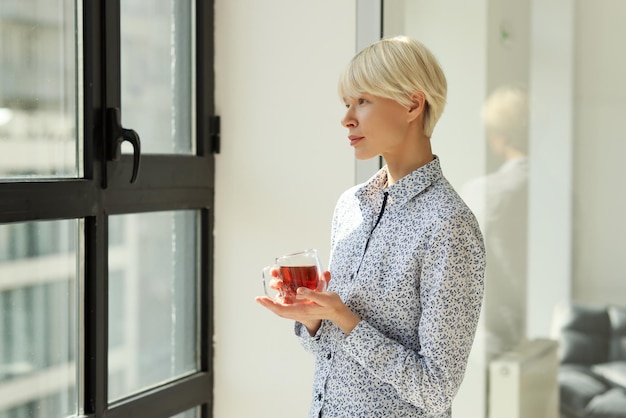 Задумчивая женщина со стеклянной чашкой чая смотрит в окно в офисе компании. Молодой менеджер