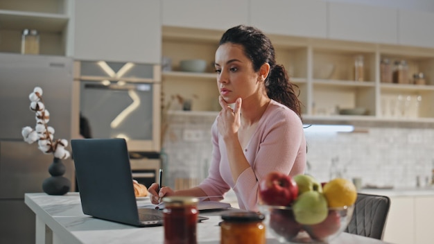 Thoughtful woman working laptop sitting kitchen close up girl making notes