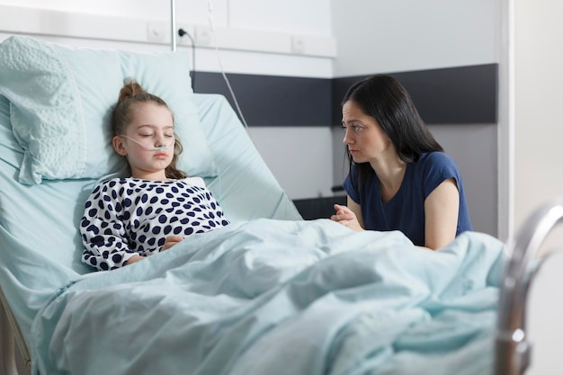 Thoughtful worried sad woman looking at sleeping ill hospitalized little girl while in pediatric hospital room. Unhappy attentive woman praying for sick daughter health condition in clinic ward.