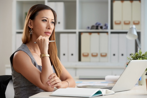 Задумчивая молодая азиатская бизнес-леди с рыжими волосами, сидящая с ручкой за столом и работающая над проектом в одиночку