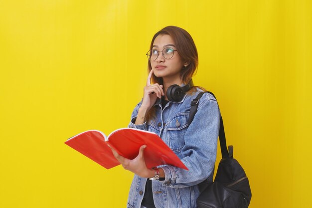 Thoughtful young asian college student standing while thinking an ideaisolated on yellow background