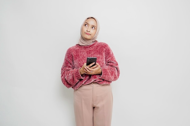 A thoughtful young Asian Muslim woman is wearing pink sweater and hijab and holding her chin isolated by white background