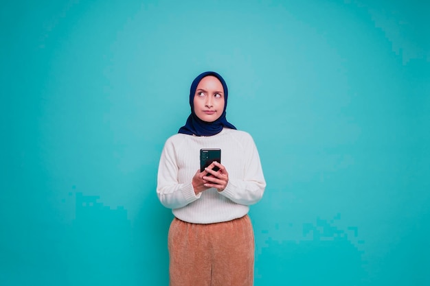 Задумчивая молодая азиатская мусульманка носит белую рубашку и хиджаб и держит подбородок на синем фоне