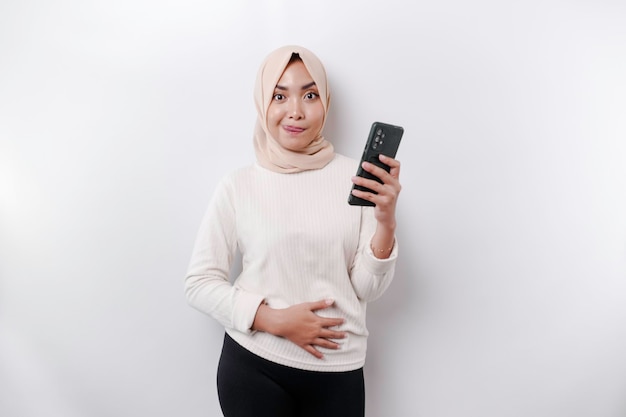 A thoughtful young Asian Muslim woman wearing a hijab and holding her chin while talking on the phone isolated by a white background