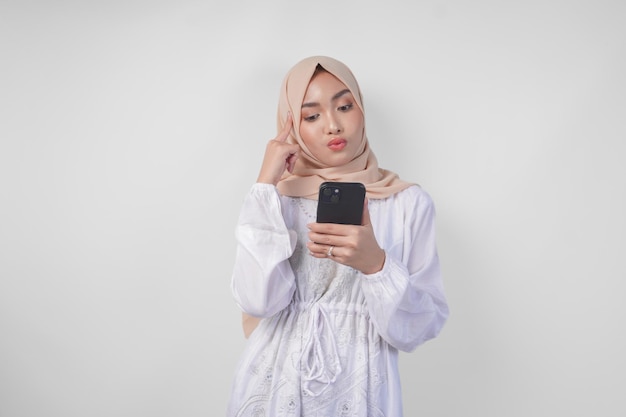 Foto giovane musulmana asiatica pensierosa che indossa un vestito bianco e un hijab che usa lo smartphone mentre tiene il mento e pensa con un'espressione seria su uno sfondo bianco isolato