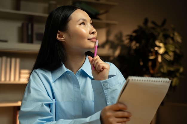 Photo thoughtful young asian woman brainstorming ideas in a home library at evening