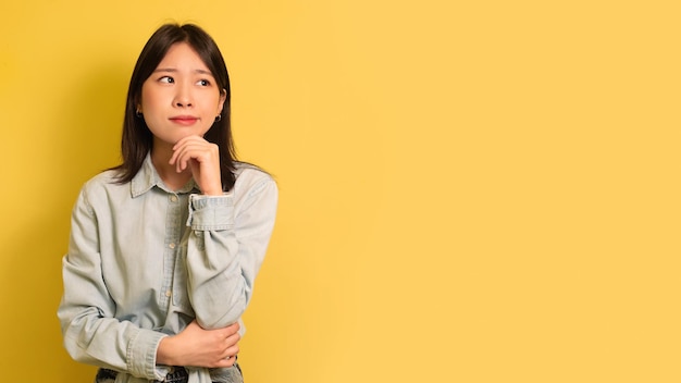 Thoughtful young asian woman in casual outfit looking at empty space over yellow studio background