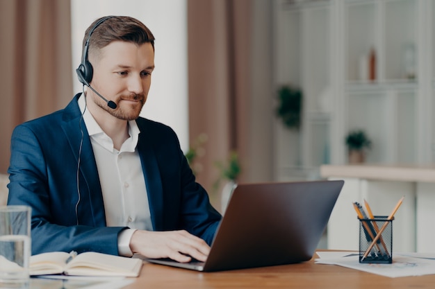 Thoughtful young bearded businessman in stylish suit making online call or taking part in web conference while using laptop and headphones with microphone. Remote work at home and business concept