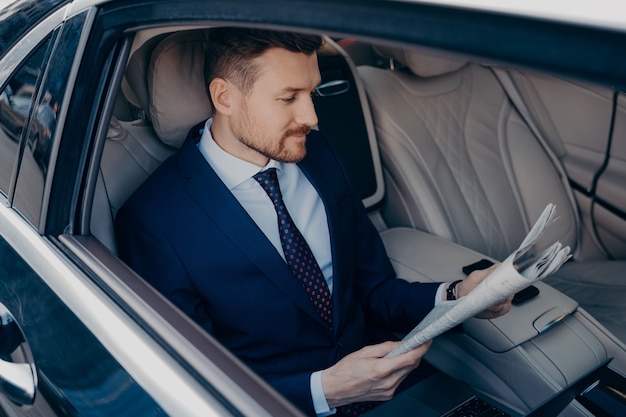 Thoughtful young bearded goodlooking executive manager in formal dark blue suit reading newspaper in backseat of limousine, rides to work at his office in business center in early morning