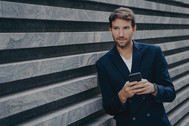 Вдумчивый молодой бородатый мужчина, стоящий снаружи, держа мобильный набор текста на клавиатуре телефона