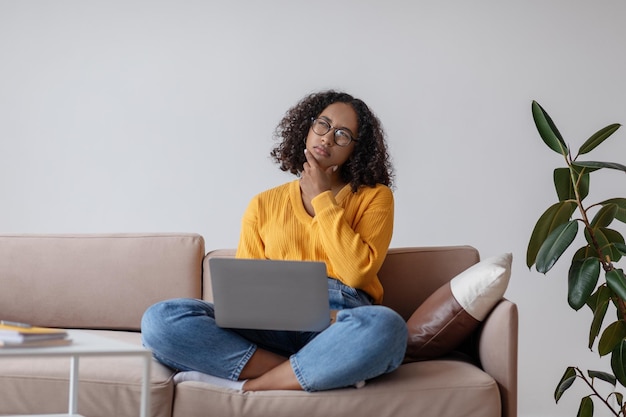 Thoughtful young black female with laptop pc sitting on couch working online from home office copy