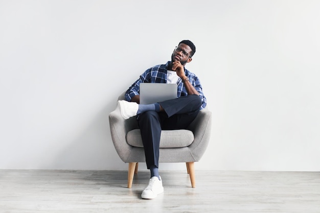 Thoughtful young black man using laptop while sitting on chair against white studio wall full length