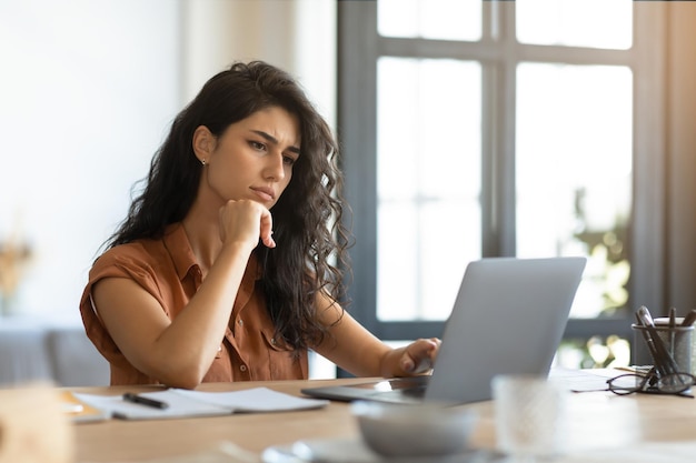 Thoughtful young brunette woman looking at laptop screen working online at desk in home office free