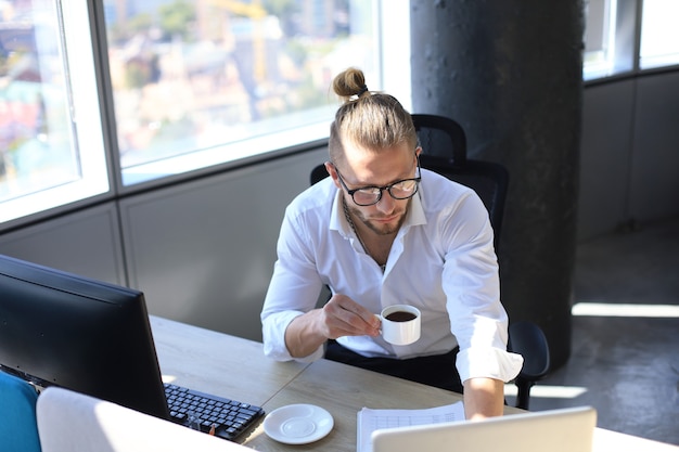 Thoughtful young business man in shirt working using computer and drinking coffee while sitting in the office.