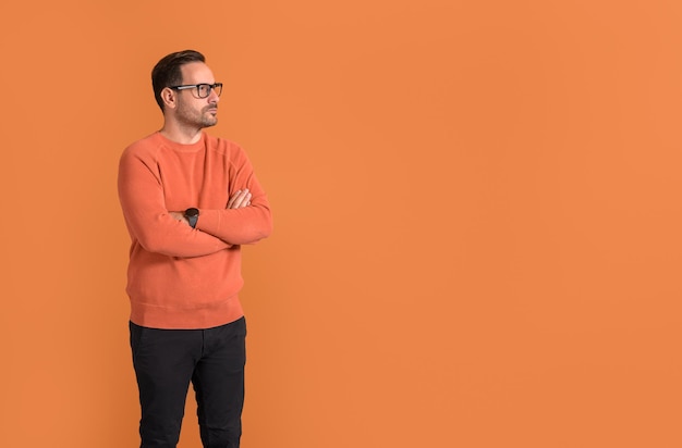 Thoughtful young businessman in eyeglasses and with arms crossed looking away on orange background