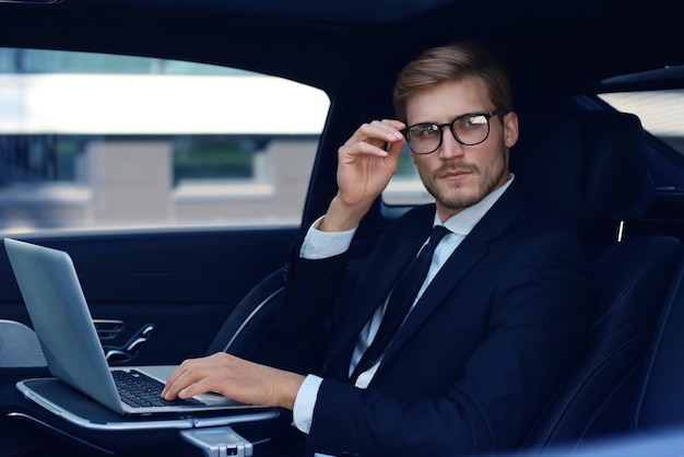 Вдумчивый молодой бизнесмен держит руку на очках, сидя в роскошной машине и используя свой ноутбук