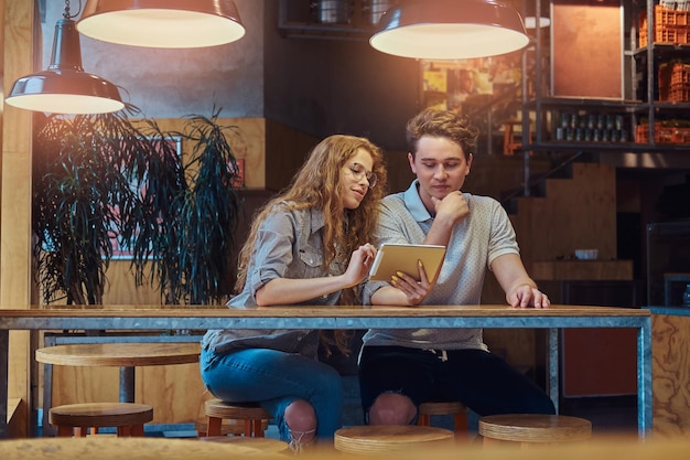 사려 깊은 젊은 부부 학생들은 휴식 시간에 대학 매점의 테이블에 앉아 있는 동안 디지털 태블릿을 사용합니다.
