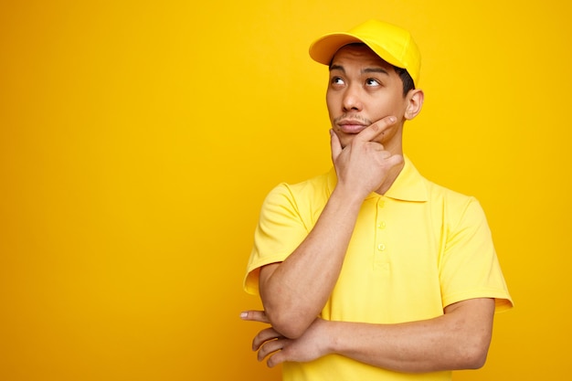 Thoughtful young delivery man wearing cap and uniform keeping hand on chin looking up 