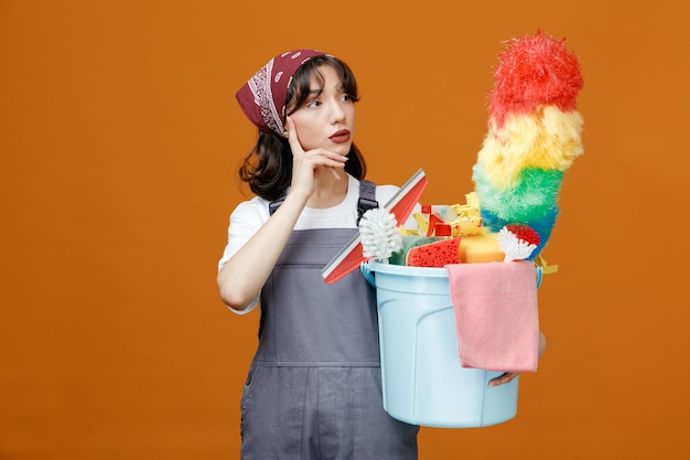 Thoughtful young female cleaner wearing uniform and bandana holding bucket of cleaning tools keeping hand on chin looking at side isolated on orange background