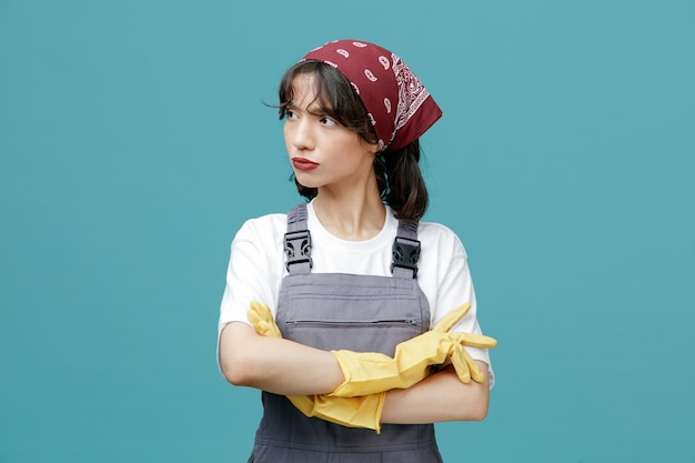 Thoughtful young female cleaner wearing uniform bandana and rubber gloves keeping arms crossed looking at side isolated on blue background