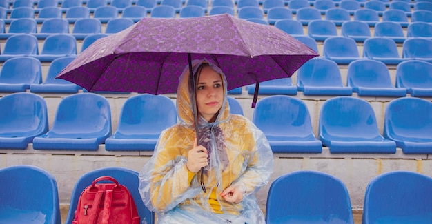 Задумчивая молодая женщина в толстовке с капюшоном и пластиковом плаще смотрит в сторону, сидя на трибуне с зонтиком в руке во время матча