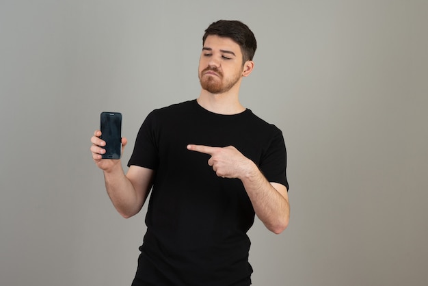 Thoughtful young guy holding phone and pointing finger at it.