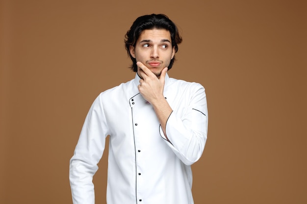 thoughtful young male chef wearing uniform keeping hand on chin looking at side isolated on brown background