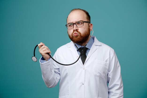 Thoughtful young male doctor wearing glasses medical coat and stethoscope grabbing stethoscope looking at side isolated on blue background