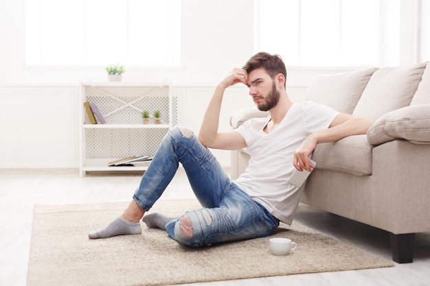 Thoughtful young man at home with mobile. Sitting on the floor with coffee. Stylish boy in casual in glasses, copy space