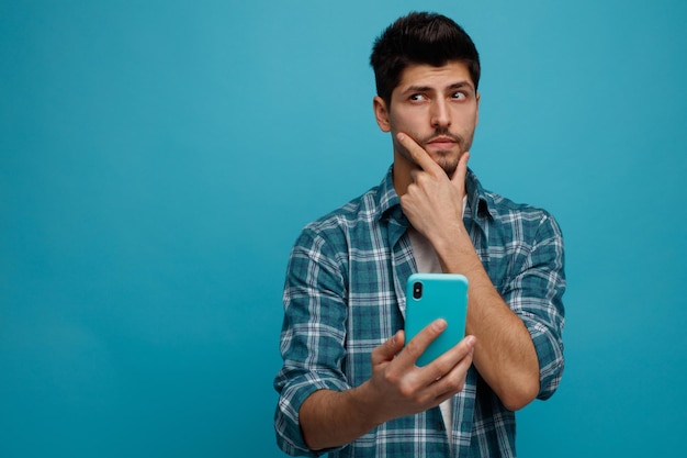 Thoughtful young man keeping hand on chin stretching mobile phone out towards camera looking at side isolated on blue background with copy space