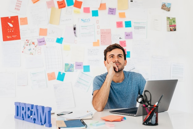 Thoughtful young man sitting near laptop against wall with notes