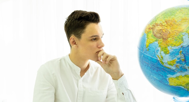A thoughtful young man in a white shirt sits at a table in front of a globe The concept of geograp