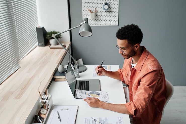 Photo thoughtful young mixed race man in eyeglasses sitting at table with lamp and analyzing graphs and ch