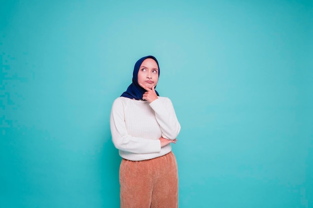 A thoughtful young muslim woman is wearing white shirt and hijab holding her chin while looking aside isolated by blue background